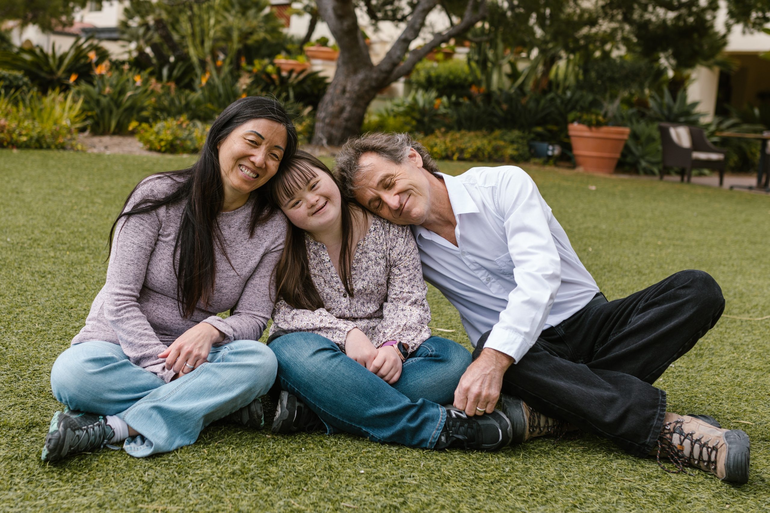 An image of parents and child with Down syndrome smiling and sitting on grass