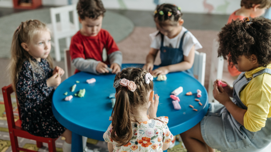 A photo of 5 children sitting at a table and playing claydough at school