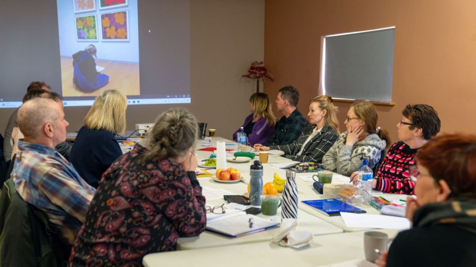 A group of people seated around a table in a workshop or meeting setting. The table is filled with notebooks, mugs, fruits, and water bottles. A projected image of a person sitting on the floor in front of colorful artwork is displayed on the wall.