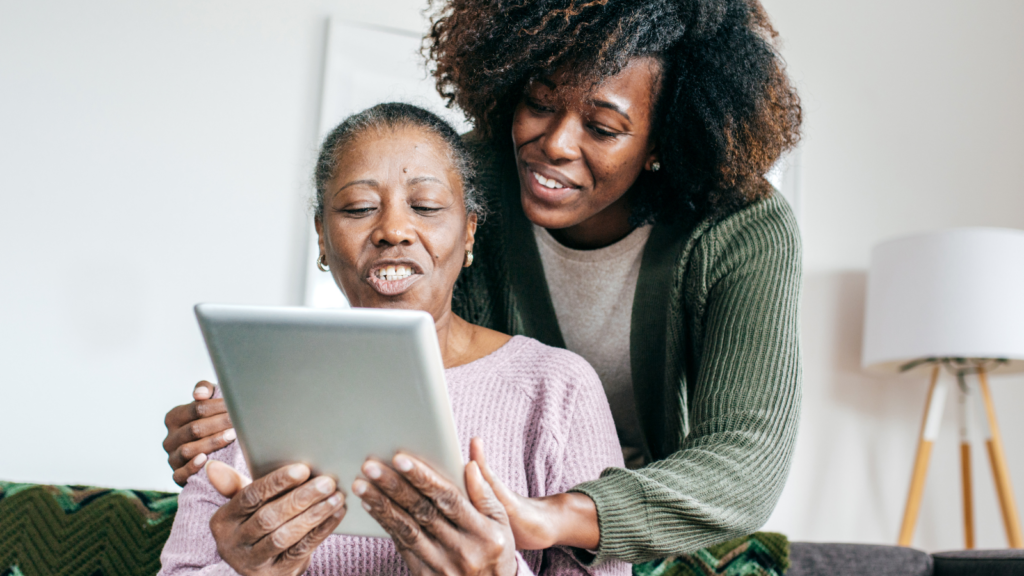 A younger woman standing behind an older woman, both smiling as they look at a digital tablet together.